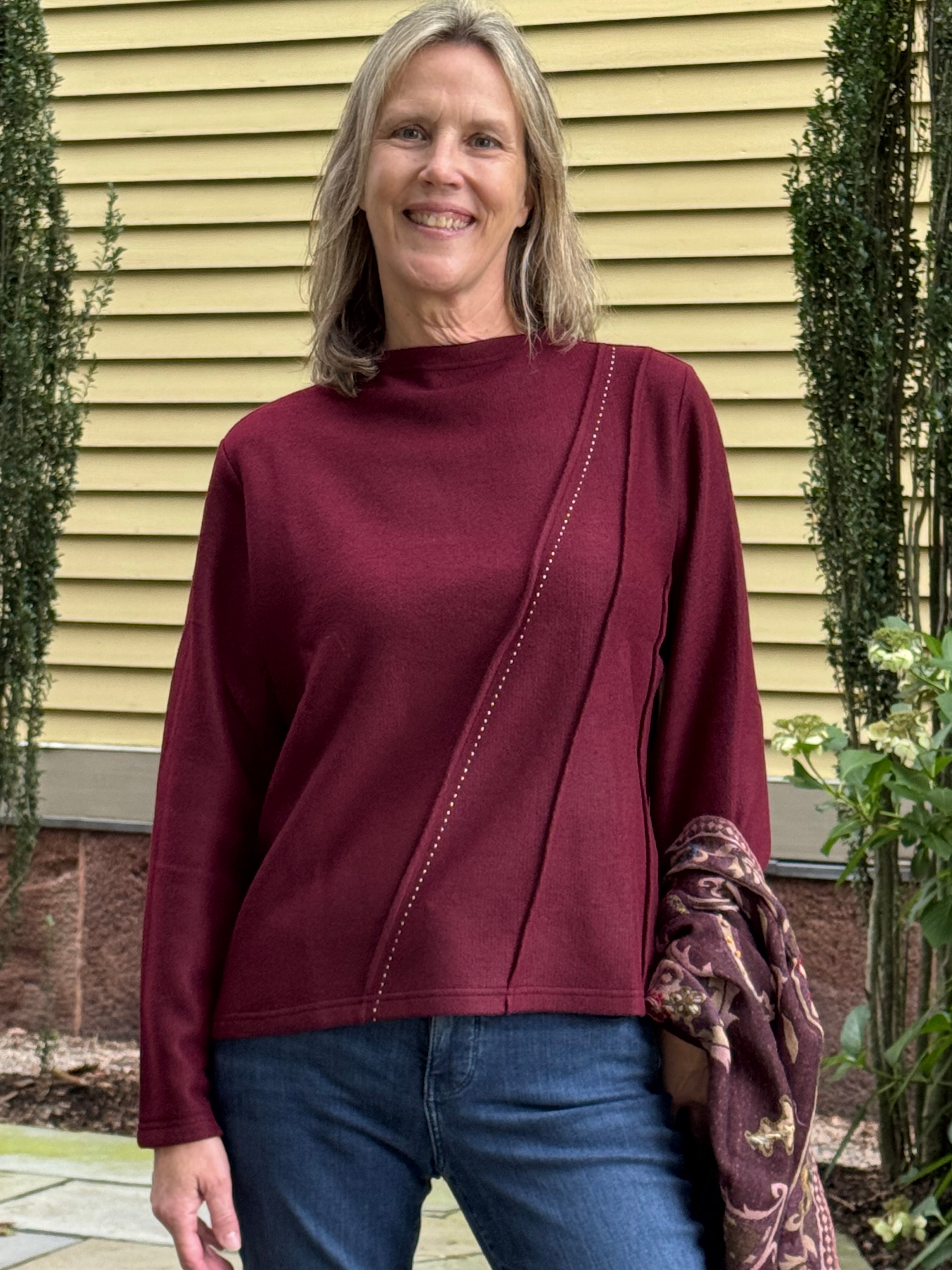 A smiling woman with blonde hair stands outdoors in front of a yellow house wall, wearing a long-sleeve mock-neck top in deep burgundy with diagonal seam detailing and blue jeans.