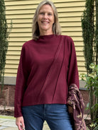 A smiling woman with blonde hair stands outdoors in front of a yellow house wall, wearing a long-sleeve mock-neck top in deep burgundy with diagonal seam detailing and blue jeans.