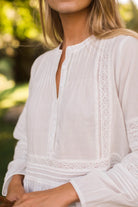 A close-up of a woman wearing a white long-sleeve blouse with delicate eyelet lace trim and a split neckline, photographed outdoors against a softly blurred green background.