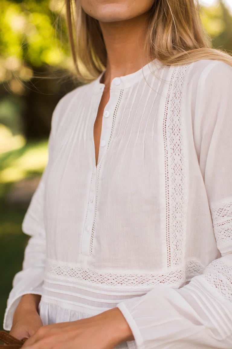 A close-up of a woman wearing a white long-sleeve blouse with delicate eyelet lace trim and a split neckline, photographed outdoors against a softly blurred green background.