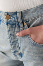 Close-up of a person wearing light blue denim jeans with a hand in the pocket, showing brass-toned metal rivets and a ribbed white top.