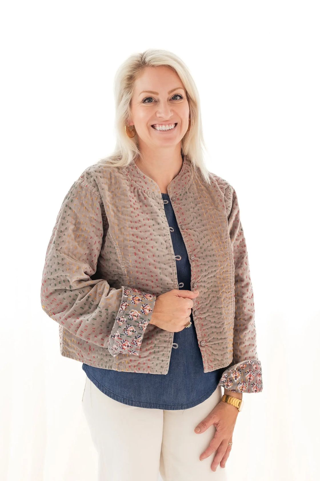 A smiling blonde woman wearing a cropped, textured beige jacket with floral-patterned cuffs over a blue button-up shirt and cream pants, photographed against a plain white background.