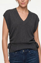 A woman wearing a dark charcoal gray sleeveless V-neck knit sweater vest with side tie details, paired with dark blue jeans, against a plain light background.