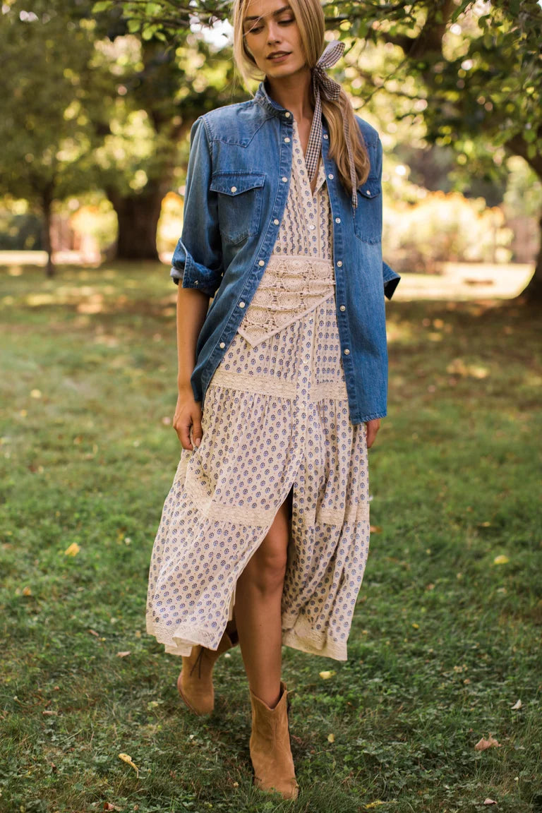 A woman standing outdoors on grass wearing a cream midi dress with a small blue floral pattern, layered under an open blue denim shirt, and wearing tan ankle boots.