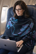 Winding River Lotus Reversible Loop Wrap in charcoal with purple and teal lotus print, worn by a woman working on a laptop on a plane