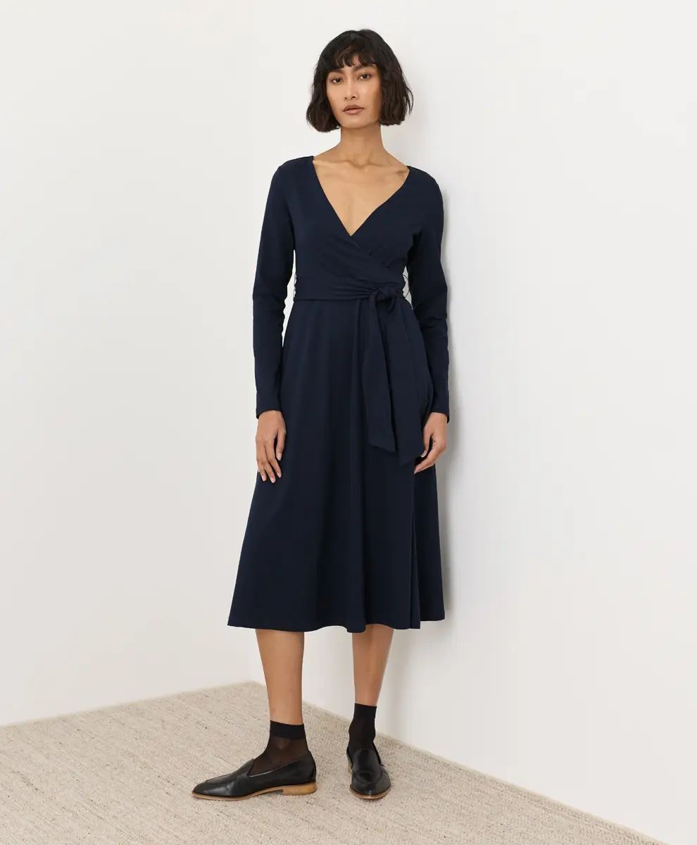 A woman modeling a dark navy long-sleeve wrap midi dress with a tie waist, paired with black socks and leather shoes, standing against a plain white wall.