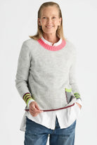 Woman wearing the Zaket & Plover Studio Pop Sweater in Lt Marl, a soft grey knit with a pink crew neck and striped cuffs, layered over a white shirt with blue jeans