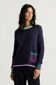 Woman wearing the Zaket & Plover Studio Pop Sweater in Navy, a relaxed crew neck knit with a green ruffled neckline, striped cuffs and a purple patch pocket
