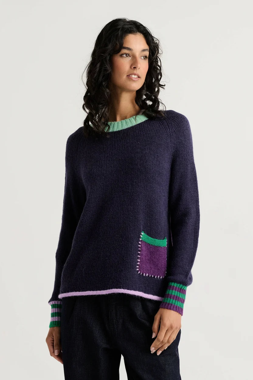 Woman wearing the Zaket & Plover Studio Pop Sweater in Navy, a relaxed crew neck knit with a green ruffled neckline, striped cuffs and a purple patch pocket